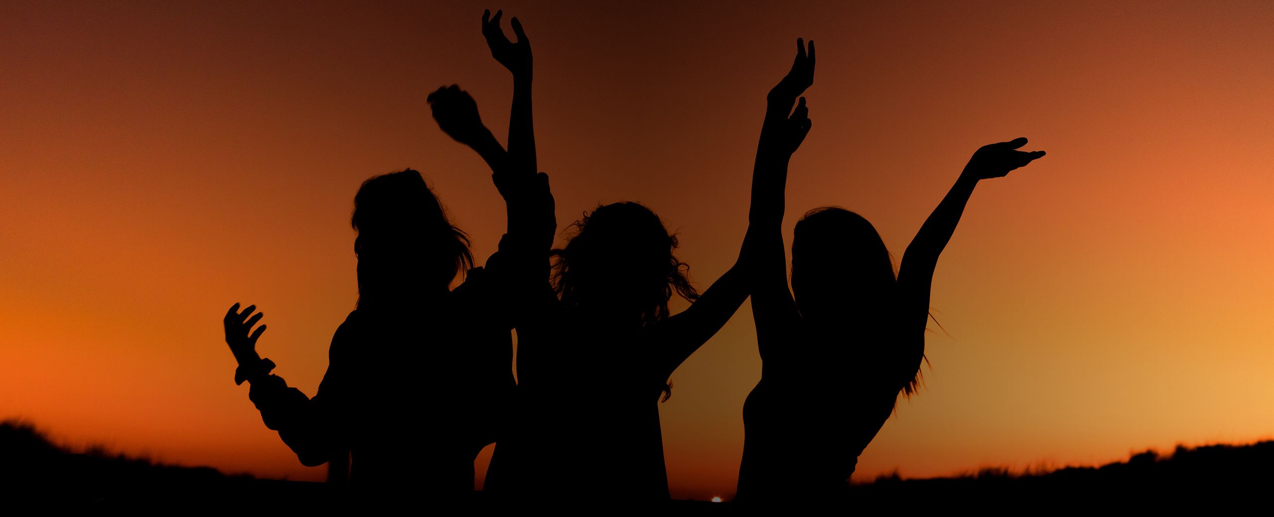Silhouette of three women dancing with hands in the air at sunset